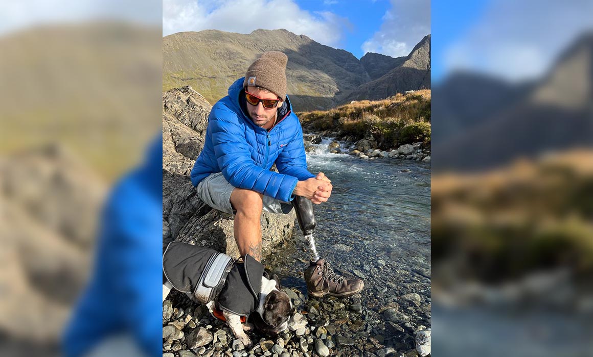 A man wearing a blue padded coat, grey shorts, brown woolly hat, and walking boots sits on a rock by some water. A small dog wearing a coat stands next to him. There are large hills in the background. The man has a prosthetic leg.