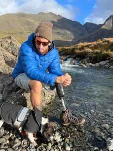 A man wearing a blue padded coat, grey shorts, brown woolly hat, and walking boots sits on a rock by some water. A small dog wearing a coat stands next to him. There are large hills in the background. The man has a prosthetic leg.