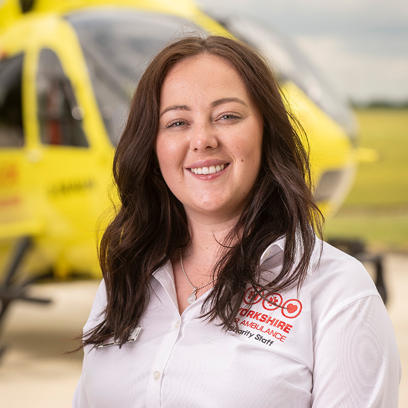 A lady with long dark brown hair, wearing a white shirt stands in front of a yellow helicopter