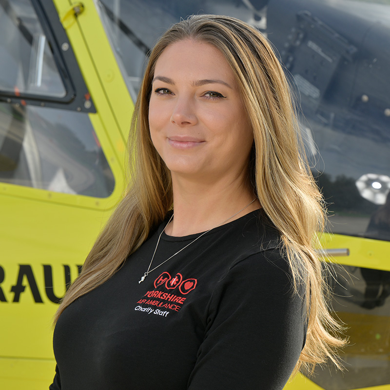 A lady with long light brown hair wearing a black top with a red logo, standing in front of a yellow helicopter.