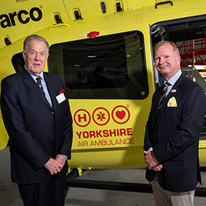 Two men wearing dark suits stand in front of a yellow helicopter