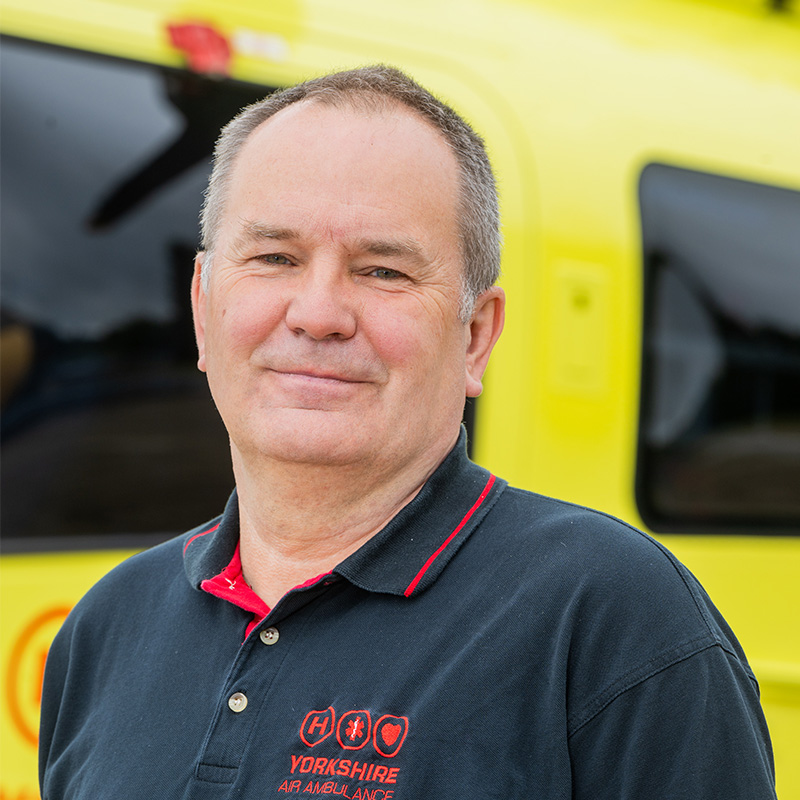Head and shoulders image of a man with short brown hair, wearing a black and red polo shirt, standing in front of a yellow helicopter.