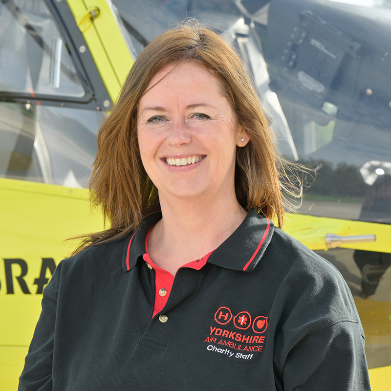 A lady with shoulder length brown hair, wearing a black t-shirt with red trim and a red logo is standing in front of a yellow helicopter.