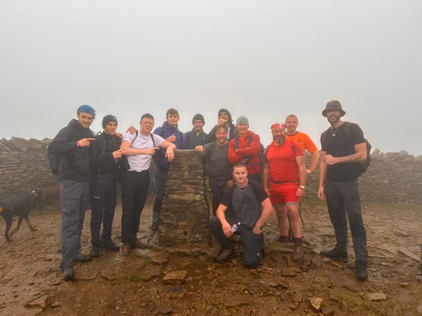 A group of men are gathering around a trig point stone with a stone wall and misty sky in the background. There is also a dog to the left of the image.