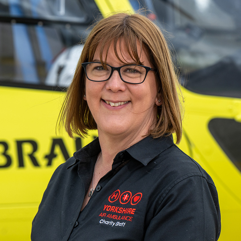 A lady with shoulder length brown hair, wearing glasses and a black shirt, with a red logo, standing in front of a yellow helicopter.