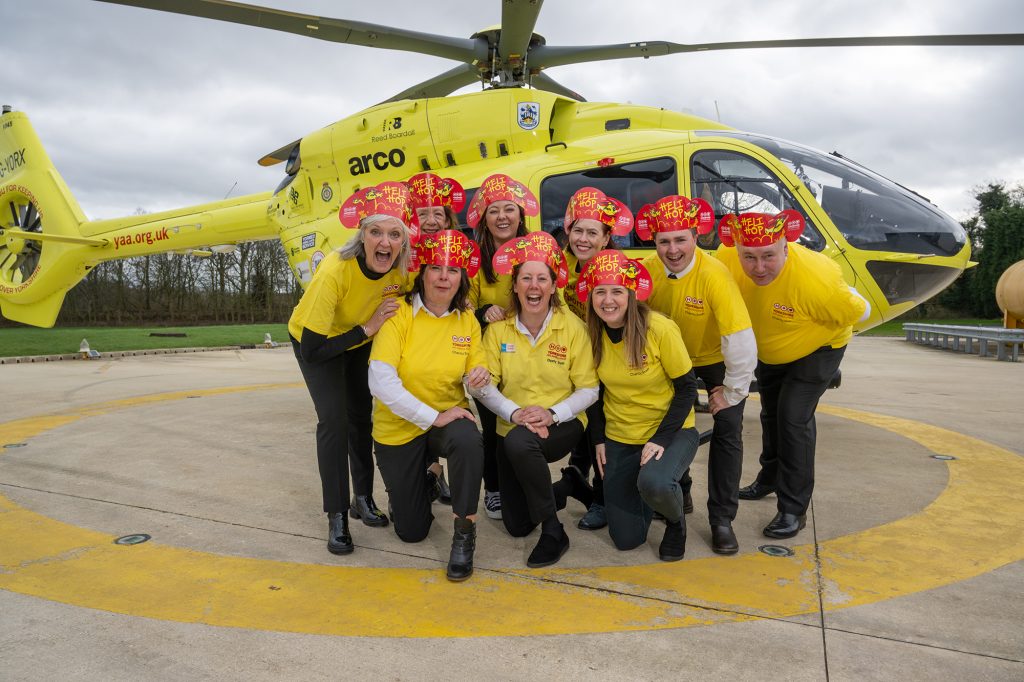 9 people wearing yellow t-shirts and red paper headbands are kneeling in front of a yellow helicopter.