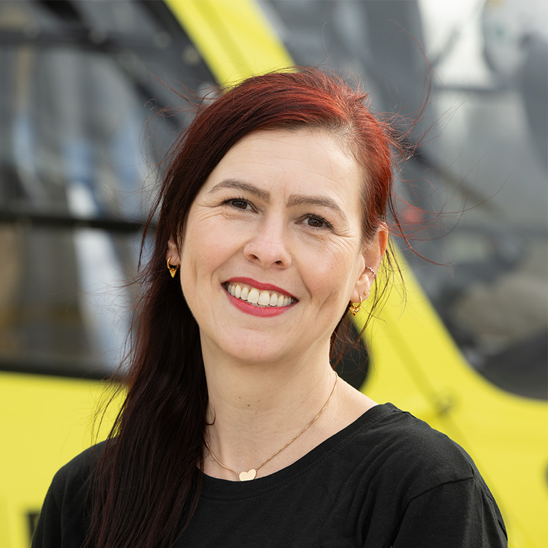 A woman with long red hair, wearing a black top with red logo and a gold heart necklace is standing in front of a yellow helicopter.