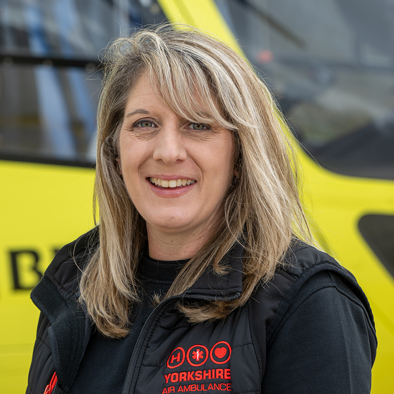 A woman with long blonde hair, wearing a black top and a black gilet with a red logo is standing in front of a yellow helicopter.