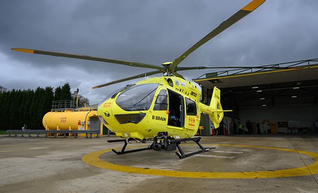 A yellow helicopter on a helipad, in front of an open hanger. There is a yellow fuel tank on the left hand side of the image.