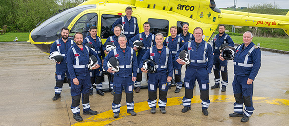 13 gentlemen wearing blue flight suits and holding white flight helmets are stood in front of a yellow helicopter.