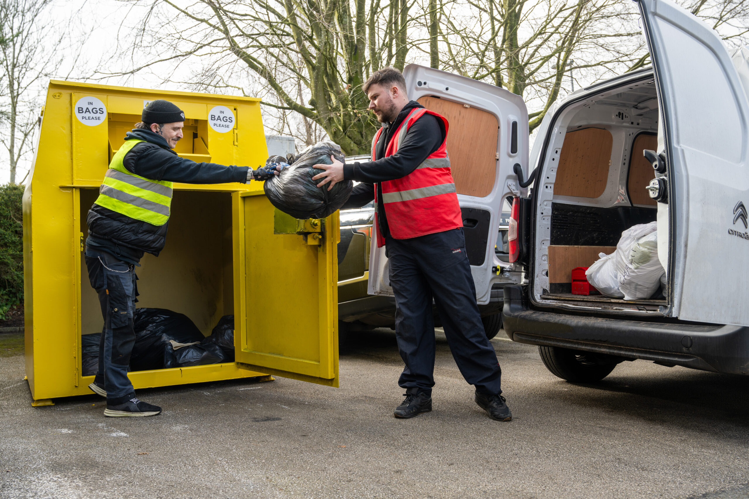 RECYCLING - EMPTYING BIN A man wearing a yellow hi vis vest is taking a bin liner from a yellow clothes recycling bin and handing it to another man, who is wearing a red hi vis vest. They are stood next to a white van, which has it's back doors open and has more bin bags in the back.