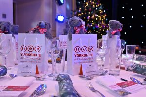 Close up of a set dinner table, with white gift bags and a lit Christmas tree in the background.
