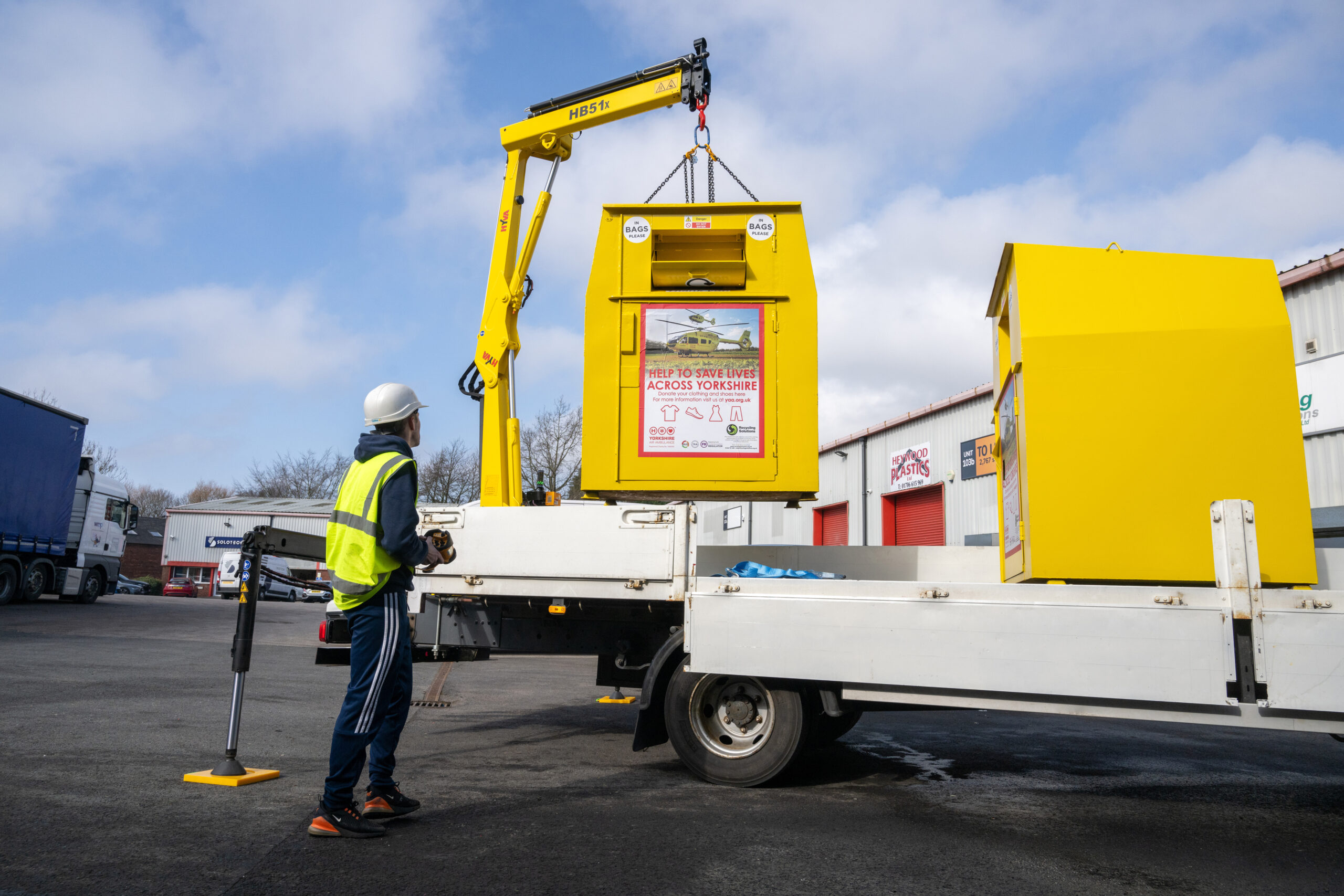 RECYCLING - HOSTING A BANK A man wearing a hi vis vest and hard hat is manoeuvring a yellow recycling bin from the back of a delivery vehicle, using a remote control.