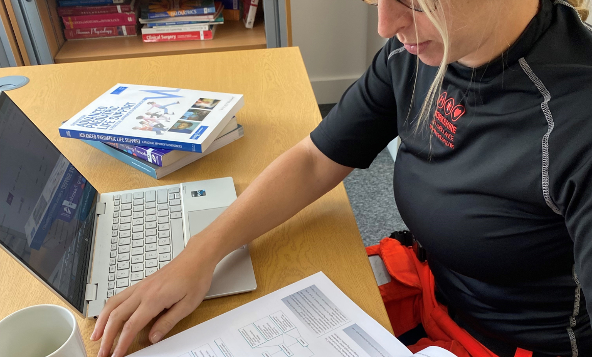 A lady wearing glasses, with blonde hair which is tied back is sat at a desk with a laptop and is reading a text book.