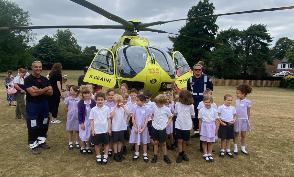 A group of school children are stood in front of a yellow helicopter in a field with two members of the helicopter crew.