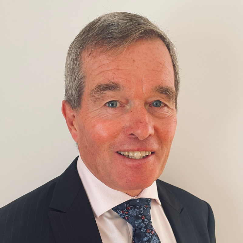 Head and shoulders image of a man with short grey/brown hair, wearing a dark coloured suit jacket, white shirt and dark patterned tie.
