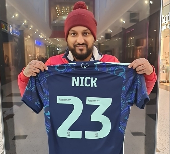 A man wearing a red bobble hat and holding a football short, with 'NICK 23' on the back.