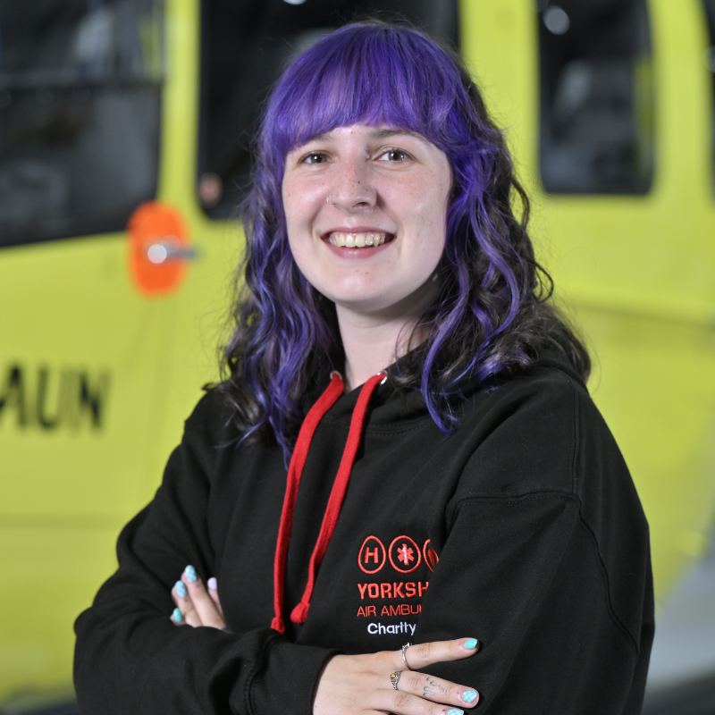 Head and shoulders photo of a person with shoulder length blue/purple hair with a fringe, wearing a black hoodie with red tassels and a red logo. They have their arms crossed and are standing in front of a yellow helicopter, only parts of which are visible.