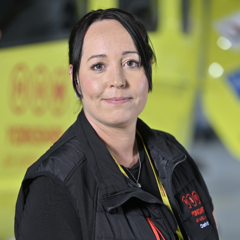 Head and shoulders photo of a person with dark hair, wearing a black top and a black gilet with red trim and a red logo. They are standing in front of a yellow helicopter, only parts of which are visible.