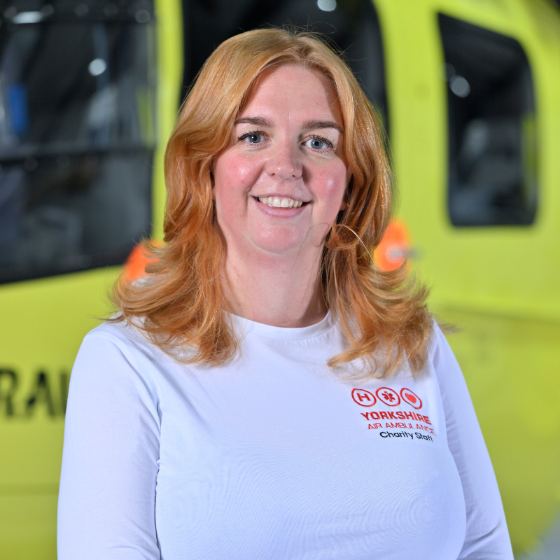 Head and shoulders photo of a person with shoulder length red hair, wearing a white top and with red logo. They are standing in front of a yellow helicopter, only parts of which are visible.