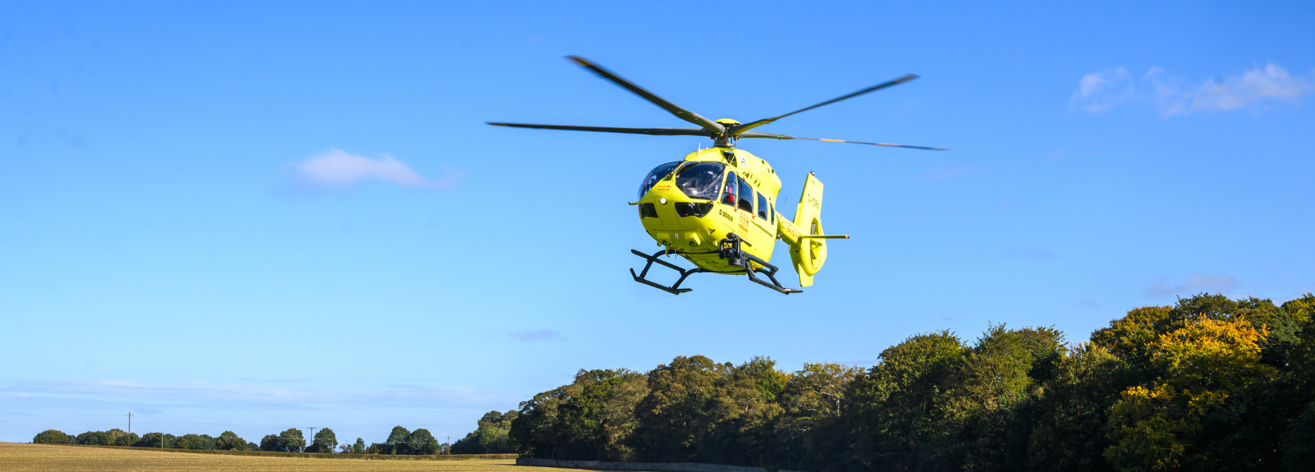 A yellow helicopter hovering in a blue sky above green trees