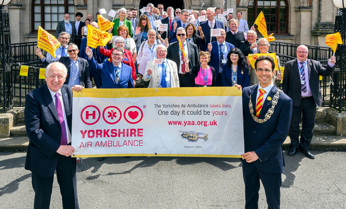 A group of people standing on the steps of an official building. Some are waving yellow or white Yorkshire Air Ambulance flags. Two people wearing suits are stood in front of the steps holding a Yorkshire Air Ambulance banner. One of those people is wearing gold chains of office.