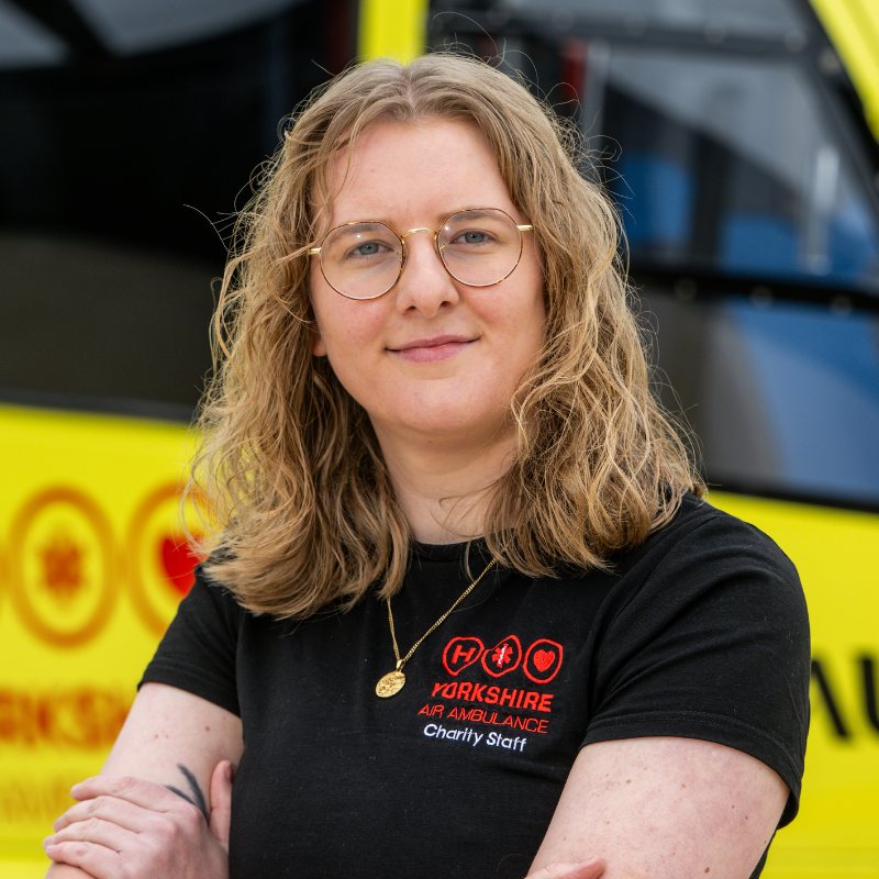 A woman with wavy blonde hair, wearing a black t-shirt with a red logo is stood in front of a yellow helicopter.