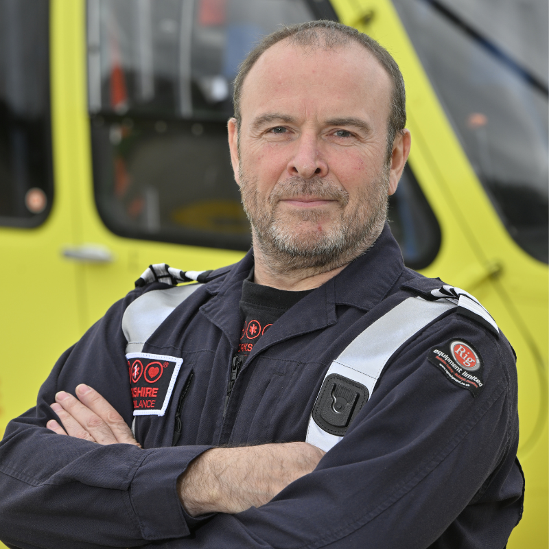 A man with short brown hair wearing a blue flight jacket is stood in front of a yellow helicopter.