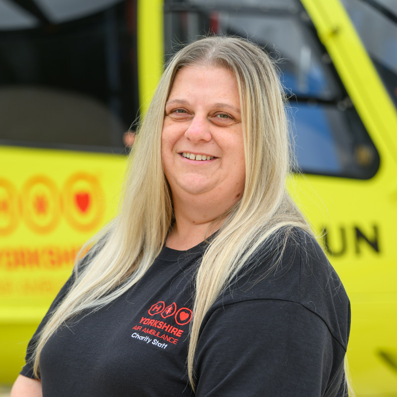 A woman with long straight blonde hair, wearing a black top with a red logo, is stood in front of a yellow helicopter.
