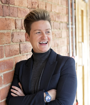 Caroline Wherritt - Rework by Respace 300 x 350 A person with short blonde/brown hair, wearing a dark suit jacket and a dark roll neck top is standing against a brick wall, with their arms crossed.