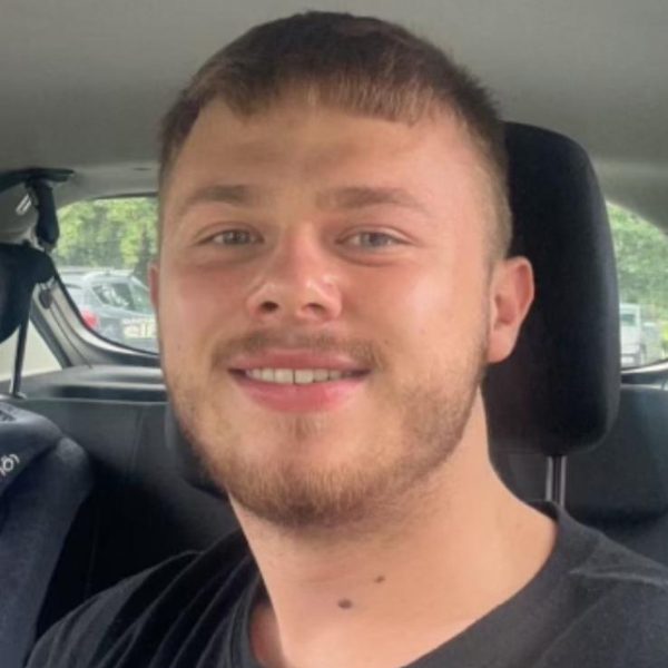 Head and shoulders image of a young man with short brown hair. He is wearing a dark brown t-shirt and is sat in a car.