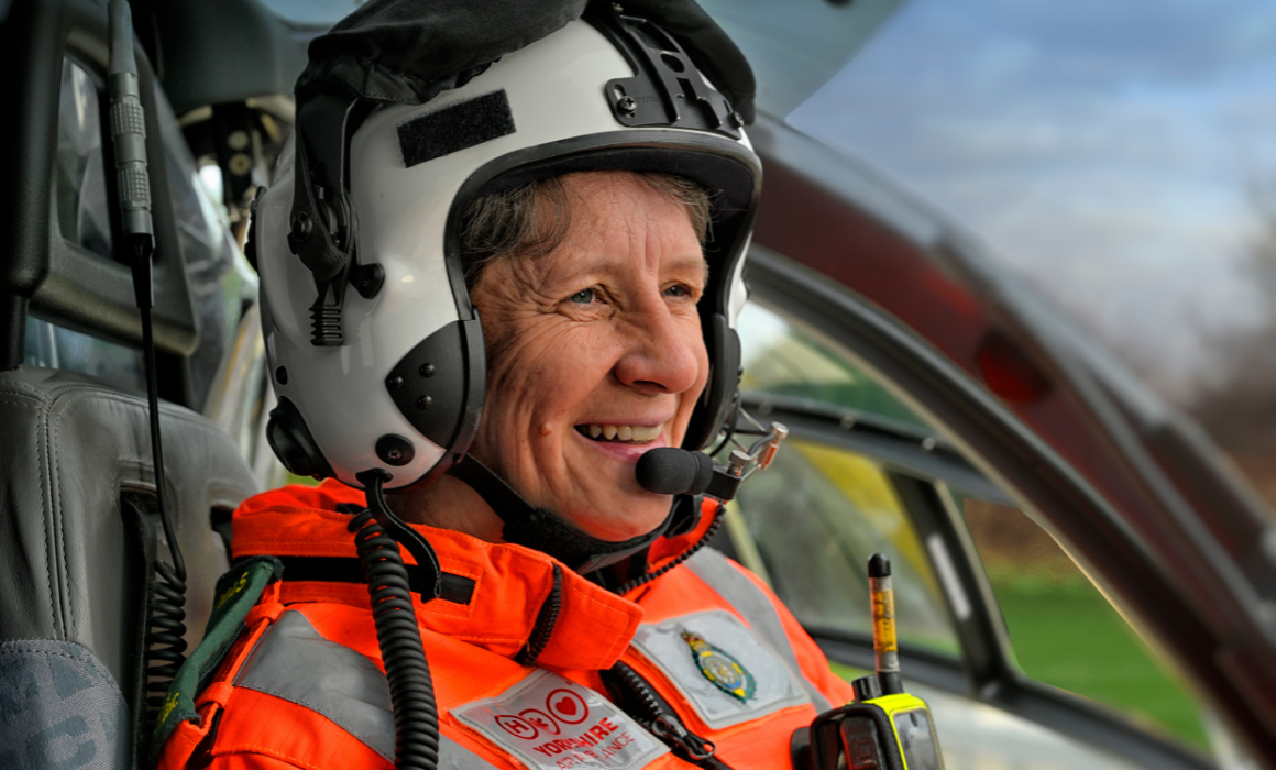 A person wearing an orange flight suite and a white flight helmet with microphone is sat in the front of a helicopter. They are smiling.