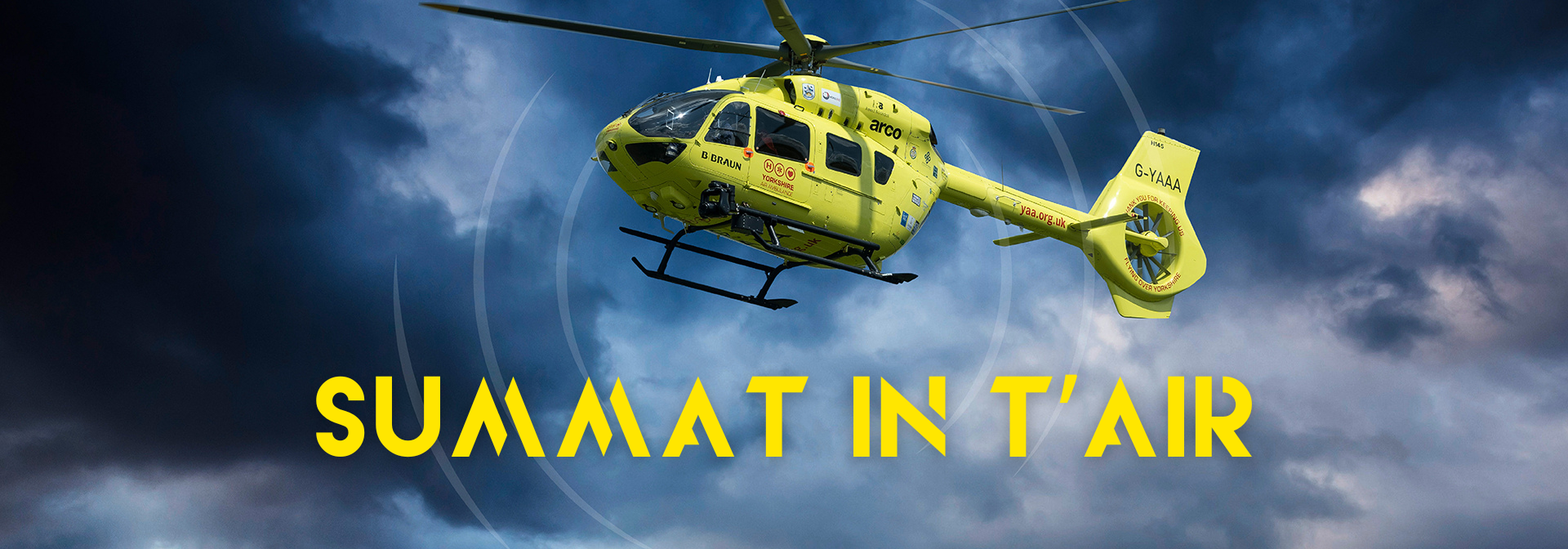 A yellow helicopter against a dark moody cloudy sky. The words 'SUMMAT IN T'AIR' in yellow.