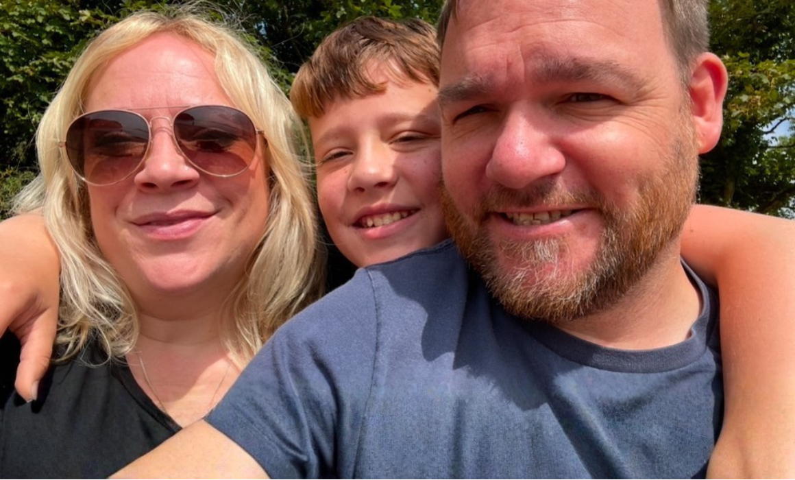 A 'selfie' image of a woman with shoulder length blonde hair, wearing sunglasses, a man with short hair and a beard, wearing a blue t-shirt and a child with short brown hair. The child's head is between the woman and the mans and he has his arms around their shoulders.