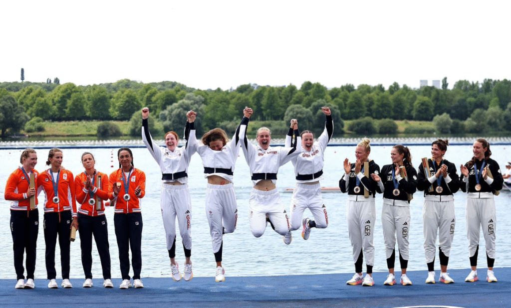 Blog Image - Podcast Ep 4 - Georgie Brayshaw Three teams, each of 4 women, all wearing team tracksuits, are standing on a platform, with water in the background. The team in the middle, wearing white tracksuits, are holding hands and jumping in the air.