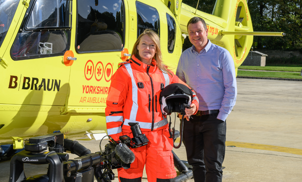 A woman with shoulder legnth blonde hair wearing an orange flight suit and holding a flight helmet under one arm and a video camera in the other, is stood next to a man with short dark hair, wearing a light blue shirt and dark coloured trousers. They are stood in front of a yellow helicopter.