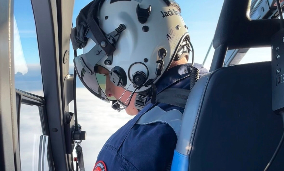 A person wearing a blue flight suit and a white flight helmet, looking out of an aicraft at clouds