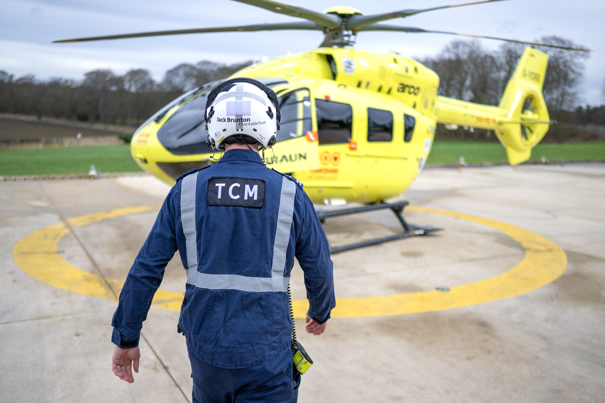 A person wearing a white flight helmet and blue flight suit, with the letters TCM on the back, is walking towards a yellow helicopter.