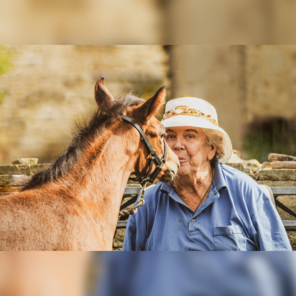 A person wearing a blue top and a cream coloured hat is kissing the nose of a light brown horse.