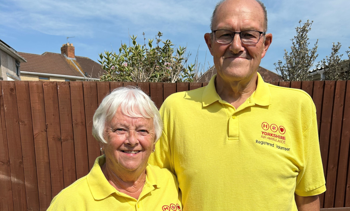 A man and woman both wearing bright yellow t-shirts which have the red Yorkshire Air Ambulance logo on and the words 'Registered Volunteer', are standing in front of a brown fence.