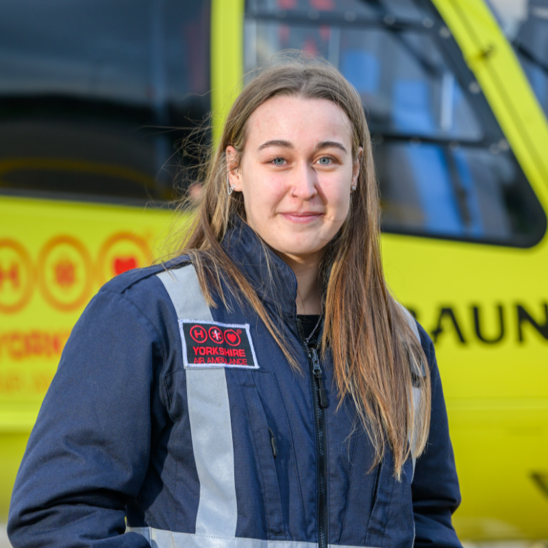 A woman with long straight brown hair wearing a blue flight jacket is stood in front of a yellow helicopter.