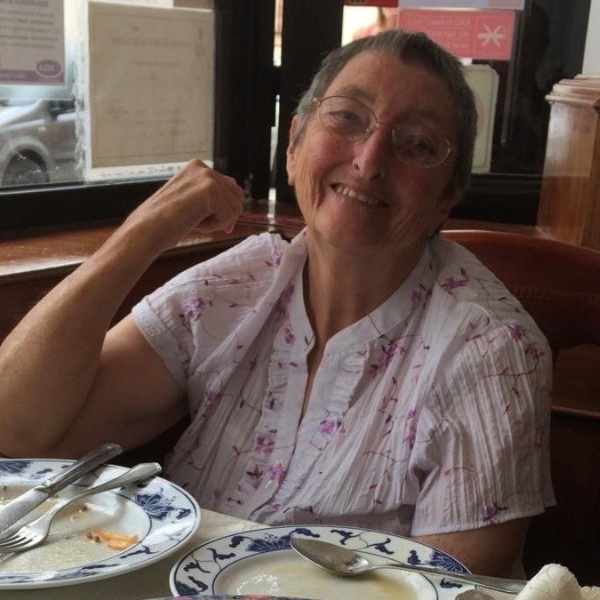A lady wearing a white short sleeved blouse with flowers on and glasses is sitting at a table smiling. She has one elbow on the table. There are empty plates on the table in front of her, it would appear she has just finished a meal.