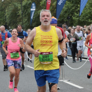 A man wearing a yellow vest with a race number pinned to it, is running along a road with other people.
