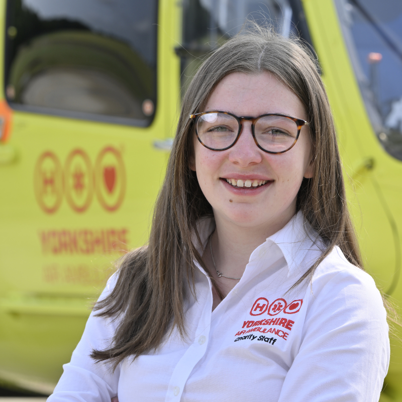 A woman with long straight brown hair, wearing black glasses and a white shirt with a red logo, is standing in front of a yellow helicopter.