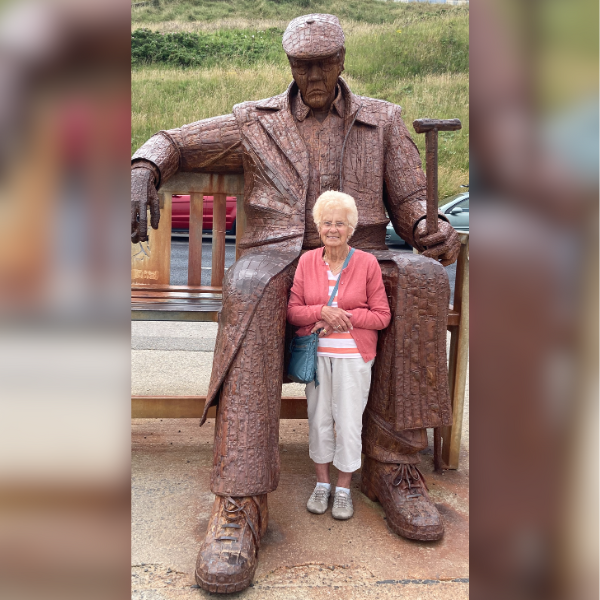 A lady with white hair is standing with a large statue of a man sat on a bench.