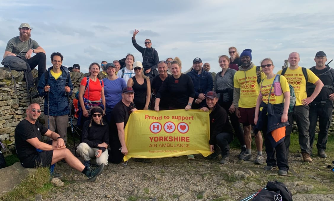 A group of people celebrating at the top of a hill, holding a yellow flag with the Yorkshire Air Ambulance logo on.