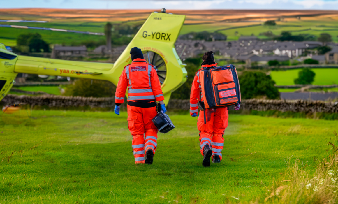 Two air ambulance crew members dressed in orange flight suits and carrying kit bags are walking towards a yellow helicopter in a field. Houses are visible in the background.