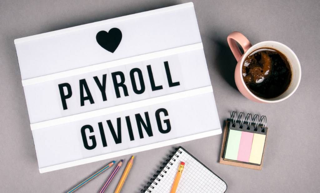 Blog Image - Payroll Giving Overhead image of a desk with a cup of coffee, some sticky notes, a note book, pens and a lightbox which says 'PAYROLL GIVING' on it.