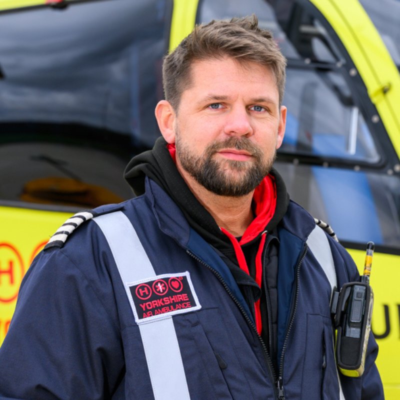 A man with short brown hair wearing a dark blue flight suit is stood in front of a yellow helicopter.