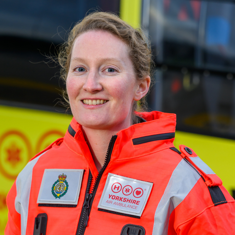 A woman wearing an orange flight suit is stood in front of a yellow helicopter.